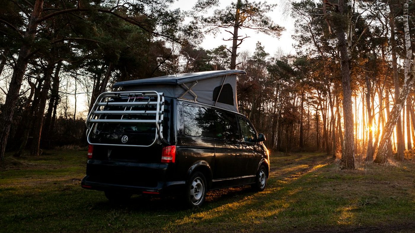 Furgoneta camper Volkswagen California negra con el techo elevable abierto en un bosque al atardecer